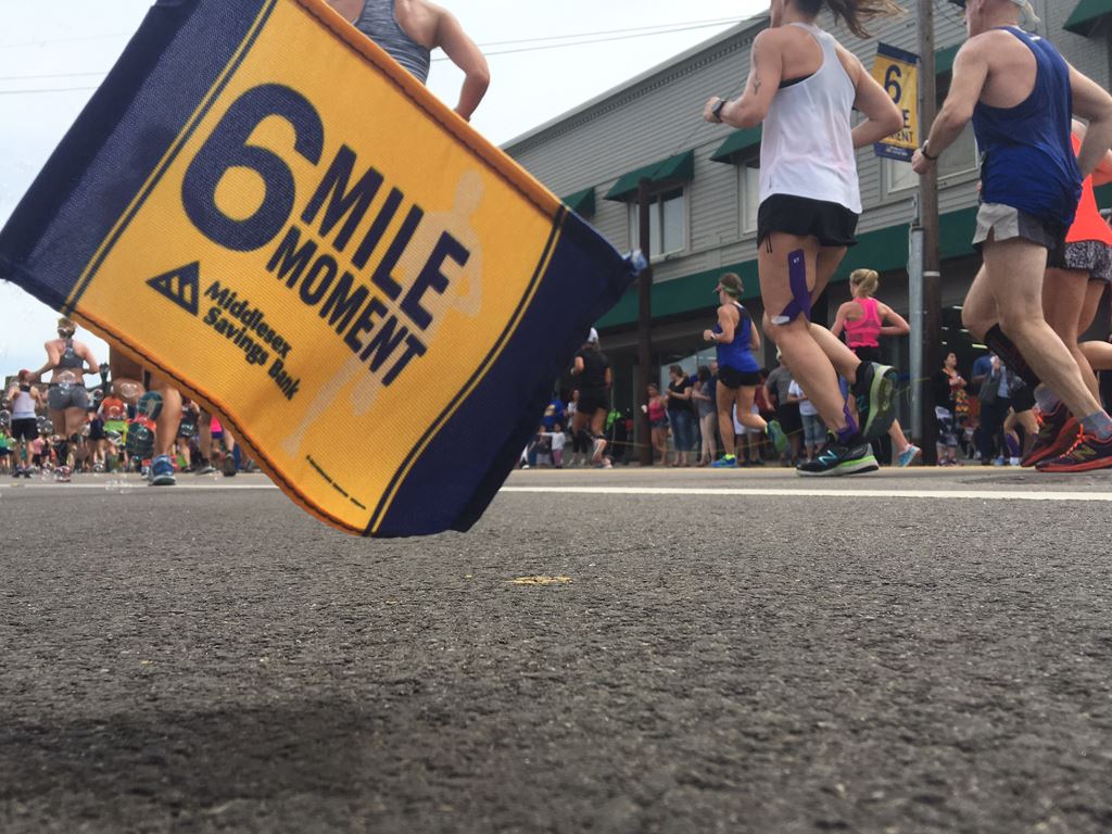 Image of 6 Mile Moment flag with runners and a building in the background from an angle at ground le